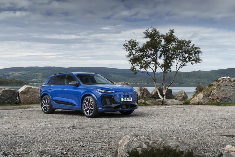 Blue Audi Q6 e-tron electric SUV parked on a lakeside gravel shore with hills behind