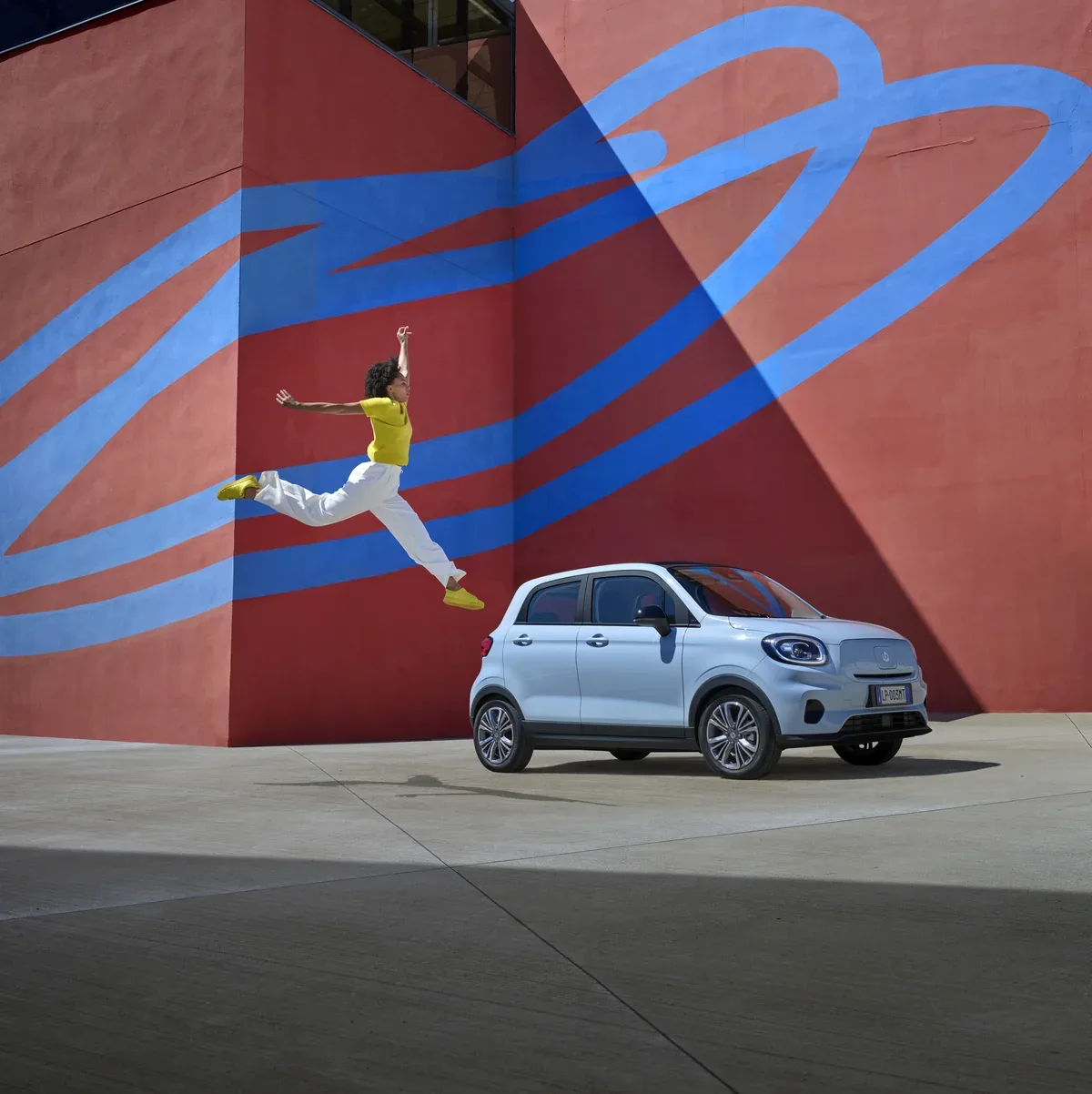 Light blue Leapmotor T03 parked by a red mural as a person jumps nearby